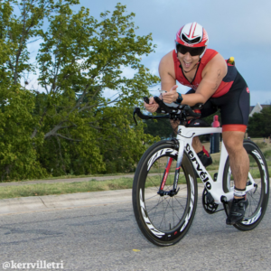 Cyclist rides in an aero position, smiling at the camera, during the 2018 Kerrville Triathlon. Credit Ed Sparks.